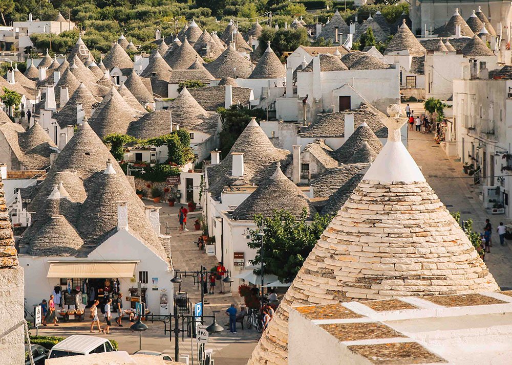 Trulli Houses in Alberobello: A panoramic view of the iconic trulli houses with conical roofs in Alberobello, Puglia, surrounded by lush greenery and bustling with tourists exploring the charming streets.
