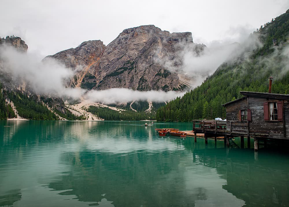 Lago di Braies in the Dolomites: A tranquil scene at Lago di Braies in the Dolomites, with misty clouds hanging over the dramatic mountain backdrop and a rustic wooden boathouse on the lake's edge, showcasing a perfect family getaway amidst nature's beauty.