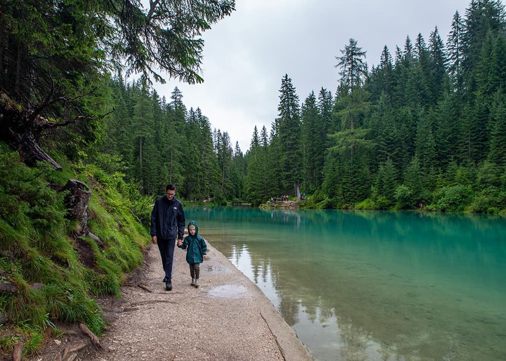 Walking by Lago di Braies: A late teen and his young brother are walking along the edge of Lago di Braies, surrounded by dense evergreen forests and the lake's stunning turquoise waters, creating a serene and family-friendly hiking experience in the Dolomites.
