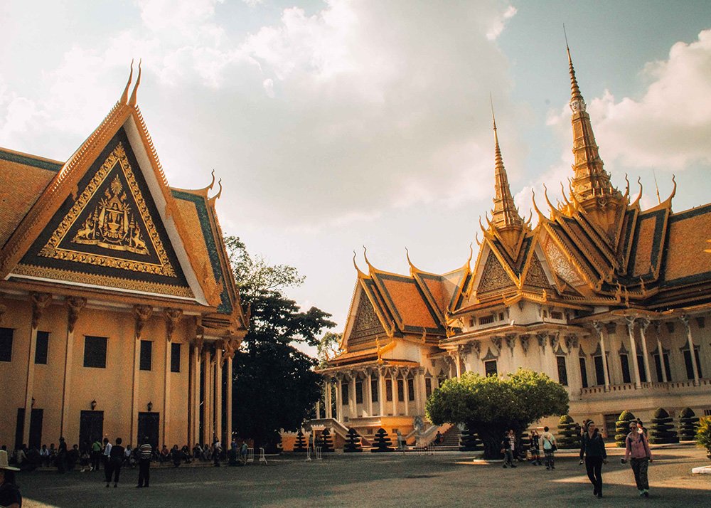 The Royal Palace in Phnom Penh, Cambodia, with its golden spires shining under a partly cloudy sky. Several tourists can be seen exploring the courtyard, highlighting the palace's stunning Khmer architecture.