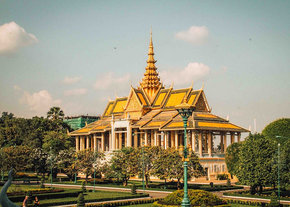 A bright and sunny day at the Throne Hall in the Royal Palace, Phnom Penh. The intricate golden roof and beautifully manicured gardens are captured, with a few visitors strolling around.
