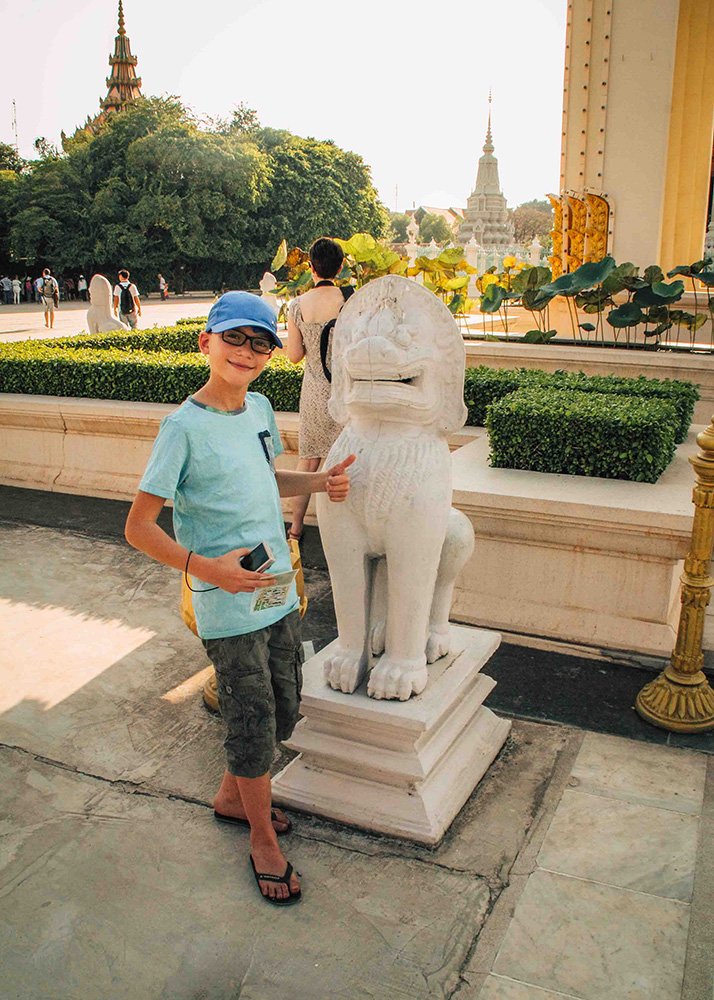 Child posing with a statue at the Royal Palace: A young boy wearing a blue cap and light blue shirt poses with a stone lion statue in the gardens of the Royal Palace in Phnom Penh. The lush greenery and temple spires in the background add to the serene atmosphere.