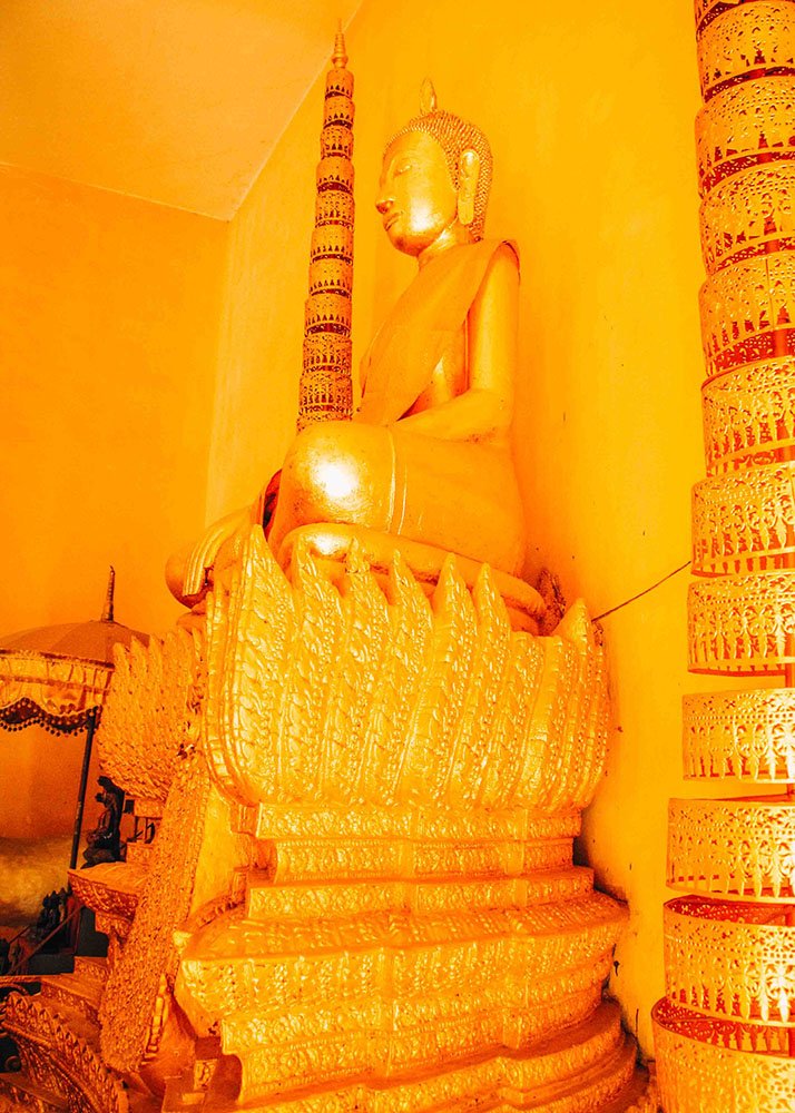 A close-up of a golden Buddha statue sitting on a lotus pedestal inside a temple in Phnom Penh. The bright yellow walls and intricate details of the statue are vividly highlighted