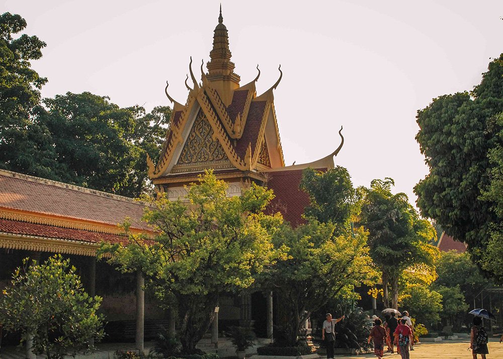 A picturesque temple with traditional Khmer architecture surrounded by lush trees in Phnom Penh. Visitors are seen walking along the shaded paths on a warm, sunny day.
