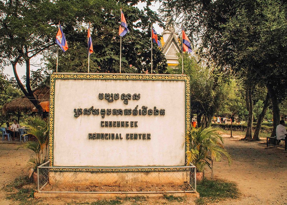 The entrance sign to the Choeung Ek Genocidal Center in Phnom Penh, with text in both Khmer and English. Cambodian flags are displayed above the sign, and visitors can be seen in the background.