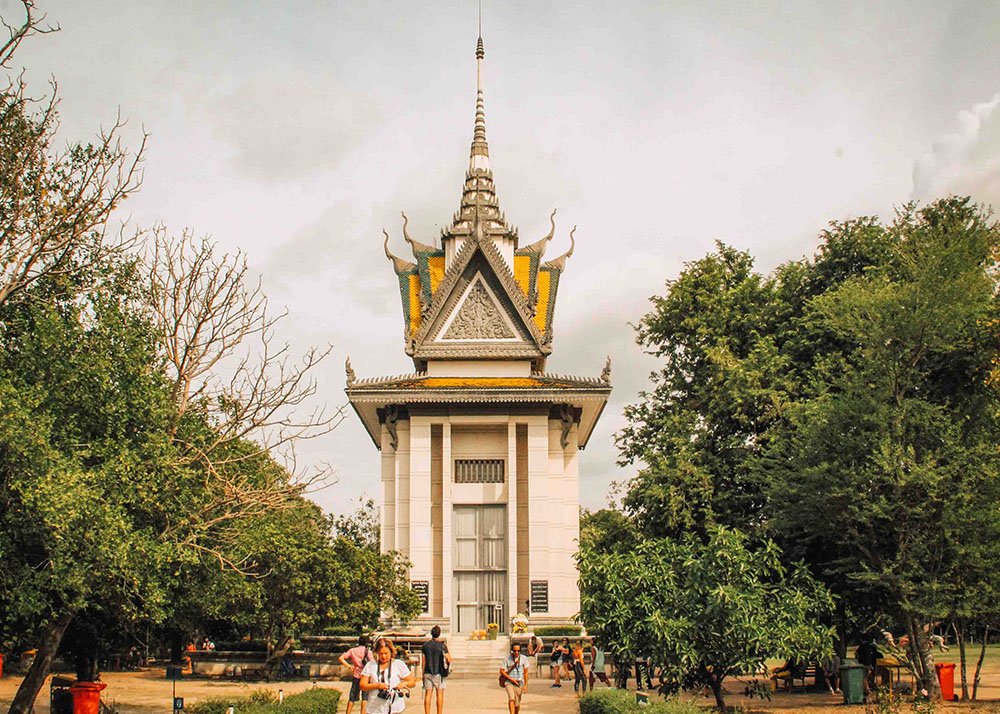 The stupa at the Choeung Ek Genocidal Center, known as the Killing Fields, near Phnom Penh, Cambodia. The memorial stands solemnly under a cloudy sky, surrounded by greenery and visitors paying their respects.