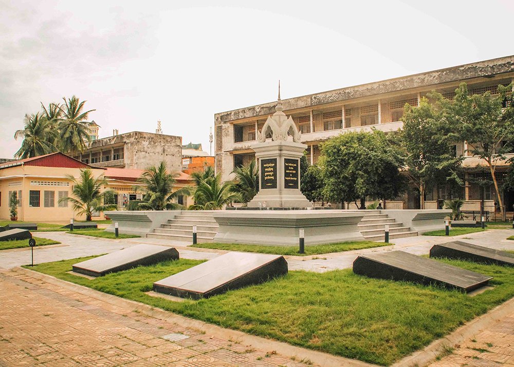 The memorial stupa at Tuol Sleng Genocide Museum in Phnom Penh, surrounded by neatly kept grounds. The scene is quiet and reflective, with the historic building in the background.