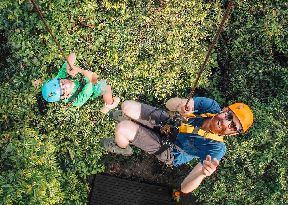 An adult and a child wearing helmets and harnesses are zip-lining through dense green foliage in the Angkor Zipline Adventure Park, Siem Reap, Cambodia. Both are smiling and looking up at the camera, with the child enjoying the adventurous activity.