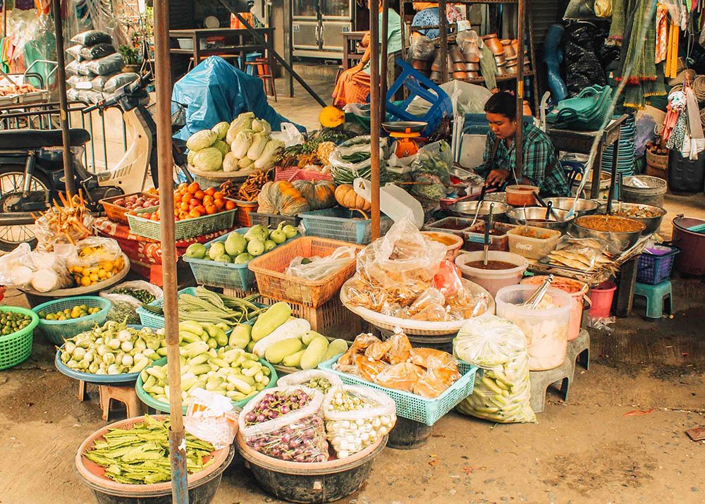 An outside market with buckets filled to the brim with fruit and vegetables. People are strolling around and buying from the ladies who sell the produce