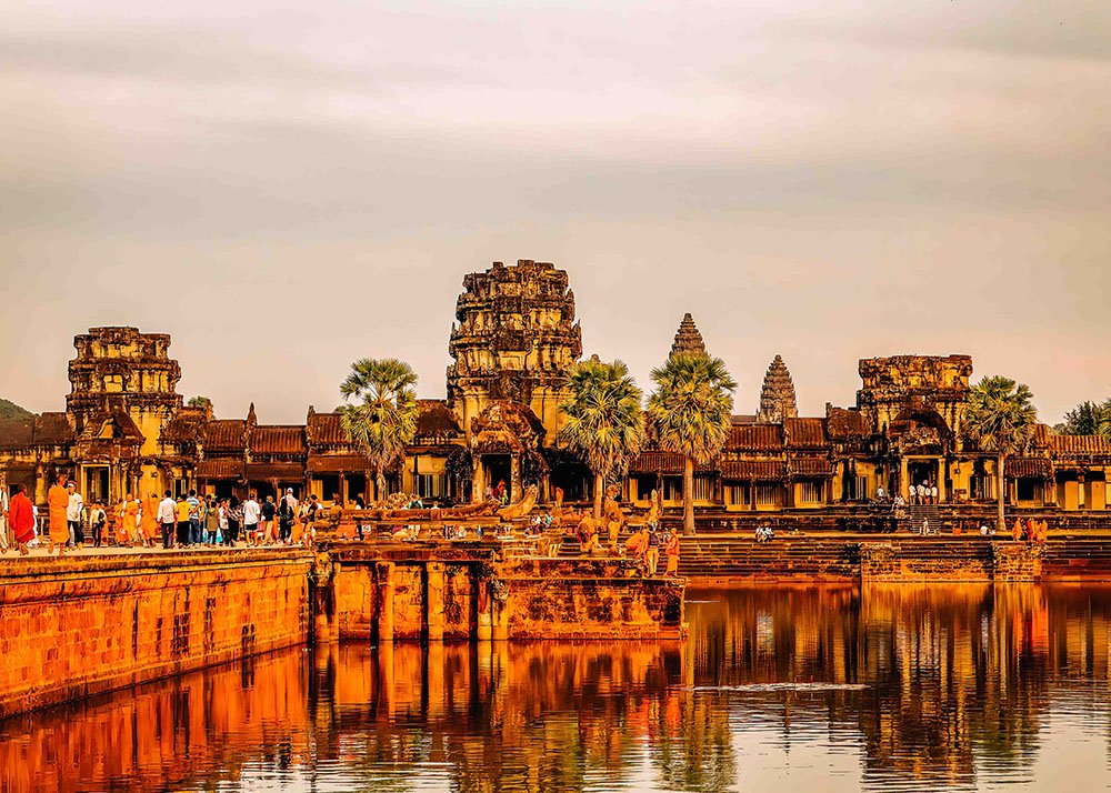 A large crowd of tourists explores Angkor Wat temple in Siem Reap, Cambodia, under a cloudy sky. The historic site is reflected in the surrounding water, enhancing its grandeur.