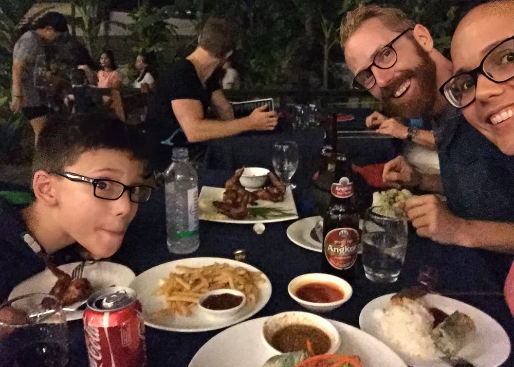 A family enjoying dinner at an outdoor restaurant in Siem Reap, featuring plates of fries, Amok, grilled meats, and a variety of dipping sauces. Angkor Beer and a Coca-Cola can are on the table, while the boy looks amused, suggesting a fun and casual dining experience during the trip.