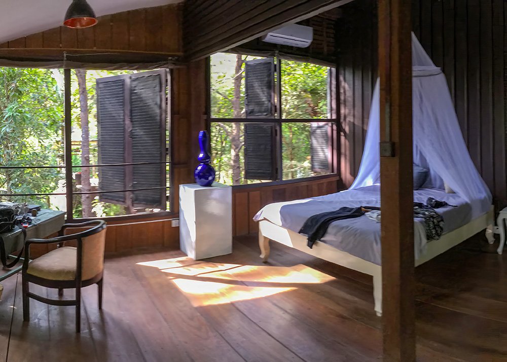 A cozy wooden cabin room at a resort in Siem Reap, featuring an open window view of lush greenery. The room has a large bed with a mosquito net and a desk near the window, providing a tranquil and rustic stay amidst the tropical landscape.