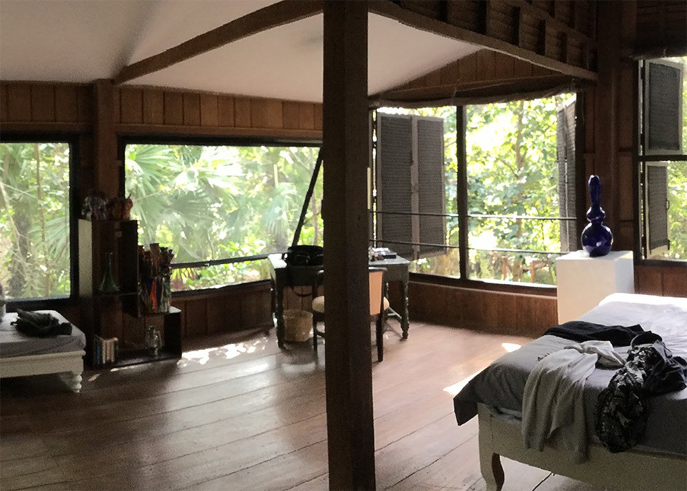 A spacious room at a Siem Reap resort with large windows overlooking a lush tropical forest. The room has a bed with a mosquito net, a desk, and a bright blue decorative vase, making it an ideal setting for a peaceful family retreat.