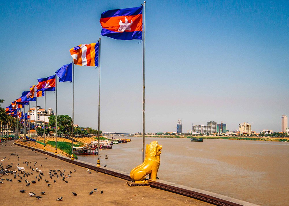 View of Sisowath Quay in Phnom Penh, Cambodia, with multiple Cambodian flags waving in the breeze along the waterfront. The quay is lined with pigeons and overlooks the Tonle Sap River, with the city skyline visible in the background on a clear, sunny day.