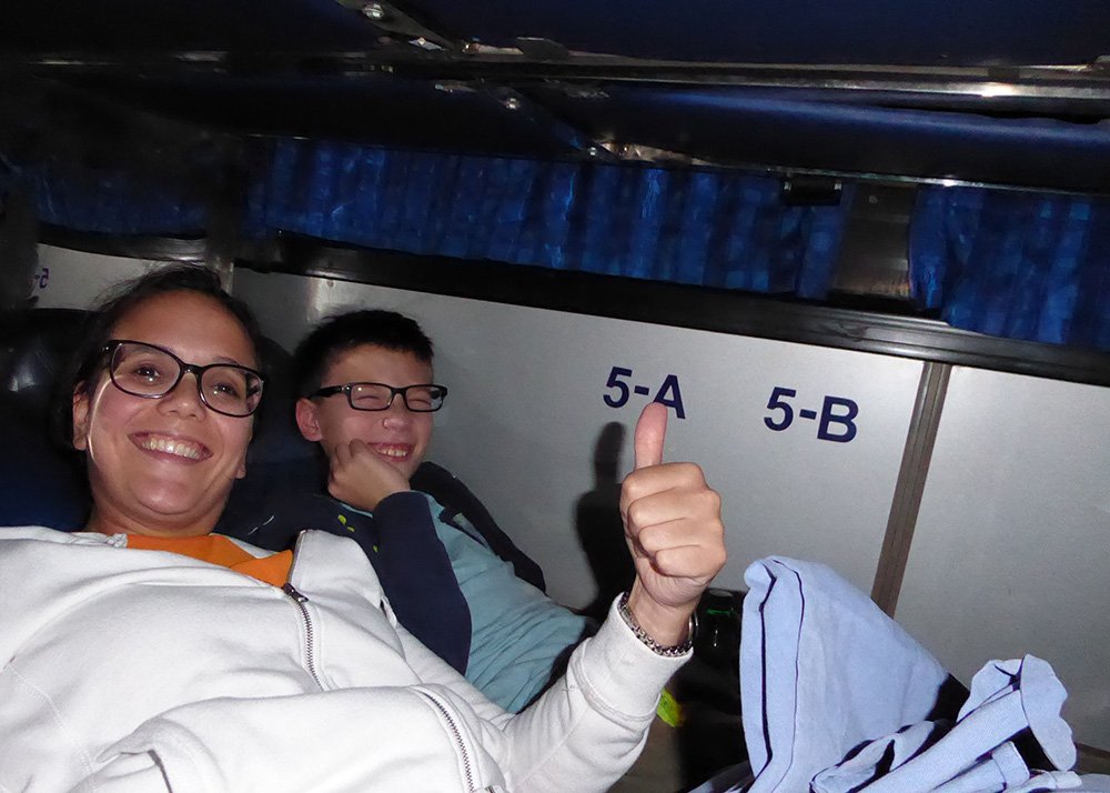 A woman and a boy are smiling inside a sleeper bus in Siem Reap, with visible seat numbers 5-A and 5-B in the background. The boy is giving a thumbs-up. Both are laying down