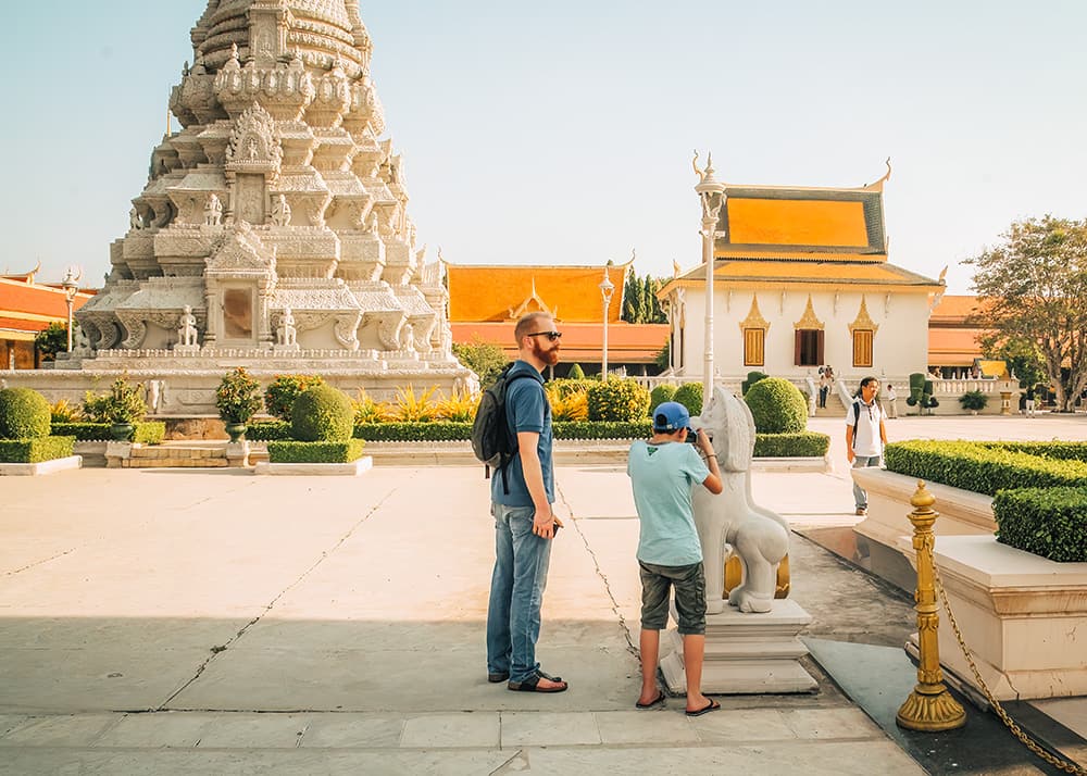 A father and son visiting the Silver Pagoda in Phnom Penh, Cambodia. The boy is examining a statue while the father looks on, with the ornate pagoda and other temple buildings visible in the background on a sunny day