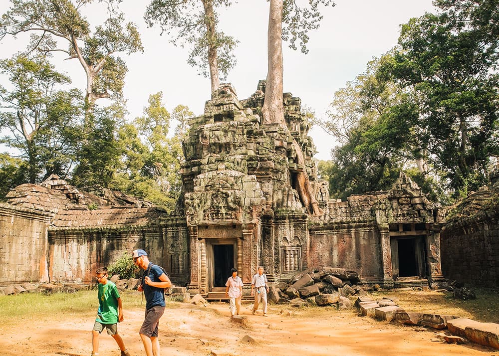 Tourists exploring the Ta Prohm temple, part of the Angkor Wat complex in Siem Reap. The ancient stone structures are intertwined with large tree roots, and the weather is sunny, ideal for sightseeing.