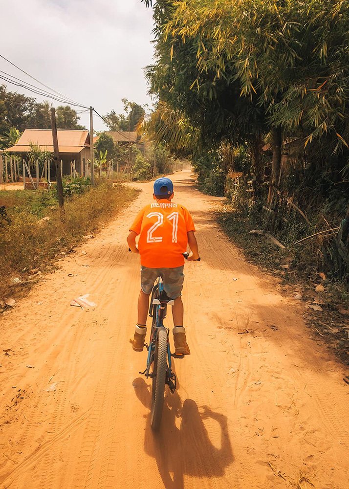 A young boy cycling along a dusty road in a rural village just outside Siem Reap in Cambodia, wearing an orange jersey with the name "Memphis" and number 21. The weather is clear, and the road is lined with bamboo and small houses, typical of Cambodian countryside.