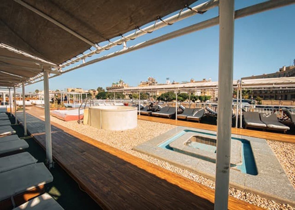 Jacuzzi and lounge area on the top deck of a cruise ship, with sunbeds and a clear view of the surrounding landscape.