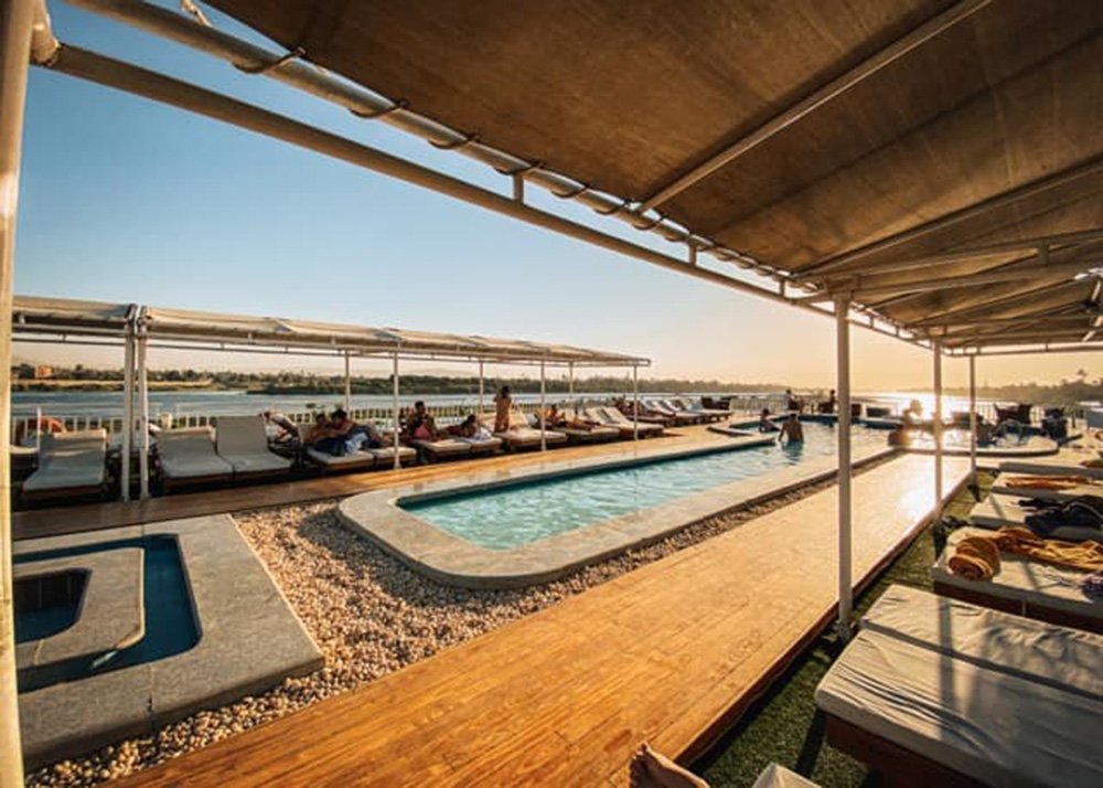 Sunlit pool deck on a cruise ship with lounge chairs, a swimming pool, and a wooden walkway, overlooking the Nile River.