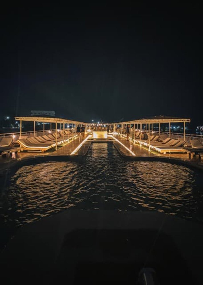 Illuminated pool deck of a cruise ship at night, with lounge chairs and a central swimming pool reflecting the lights.