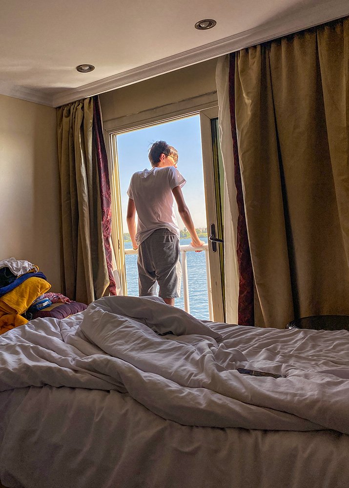 A child standing on the balcony of a cruise ship cabin, looking out over the Nile River, with an unmade bed and curtains in the foreground.