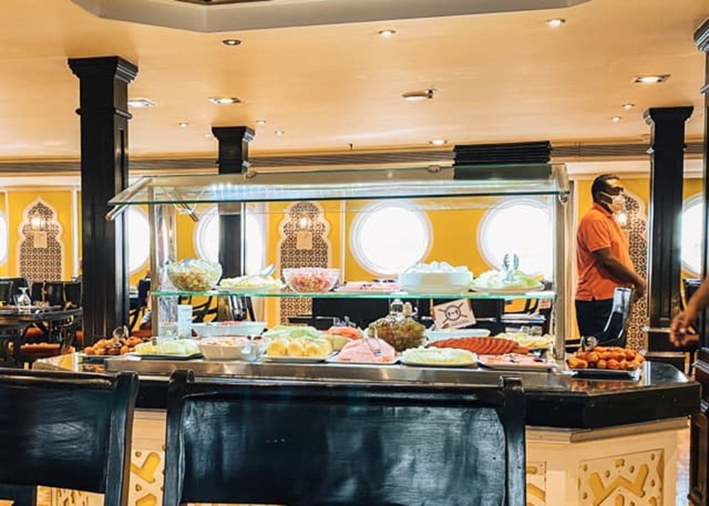 Buffet dining area on a cruise ship with a variety of foods displayed under glass, with staff and circular windows in the background.