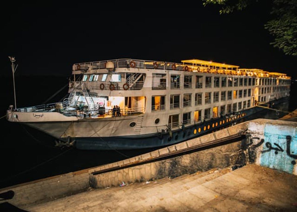 Cruise ship docked at night, illuminated by lights, with a view of the side of the ship and the dock.
