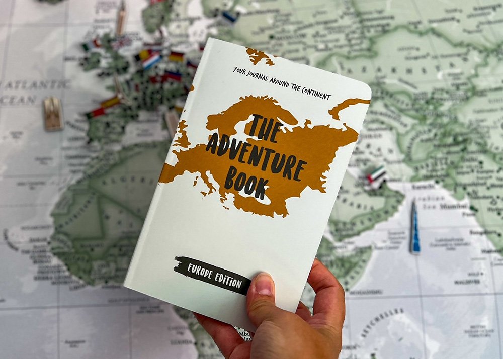 A hand holding "The Adventure Book" Europe Edition, with a white cover and a gold map of Europe, against a blurred map background. The text "Your Journal Around The Continent" is visible on the book's cover.