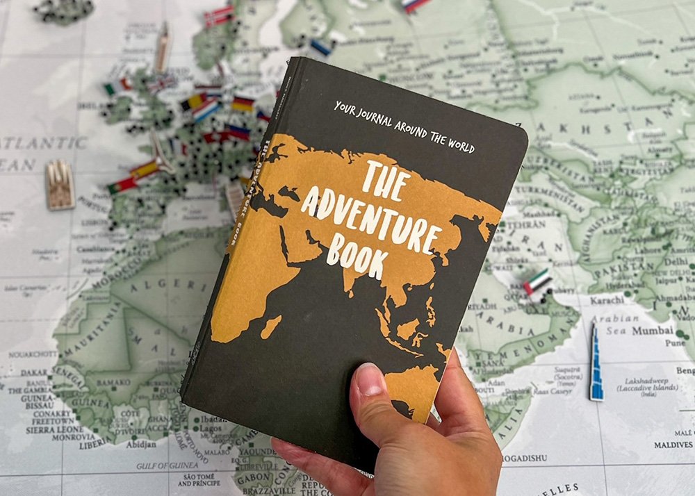 A hand holding the original "The Adventure Book" with a black cover and a gold map of the world, against a detailed map background featuring various country flags.