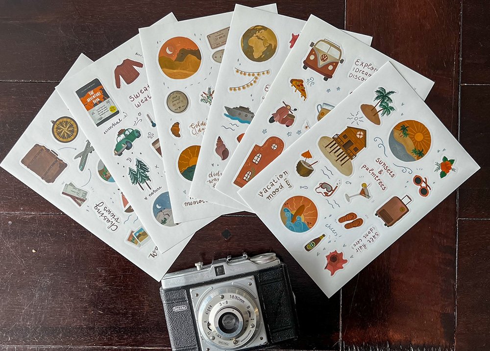 A collection of travel-themed stickers and an old school camera underneath them.The items are arranged on a dark surface.