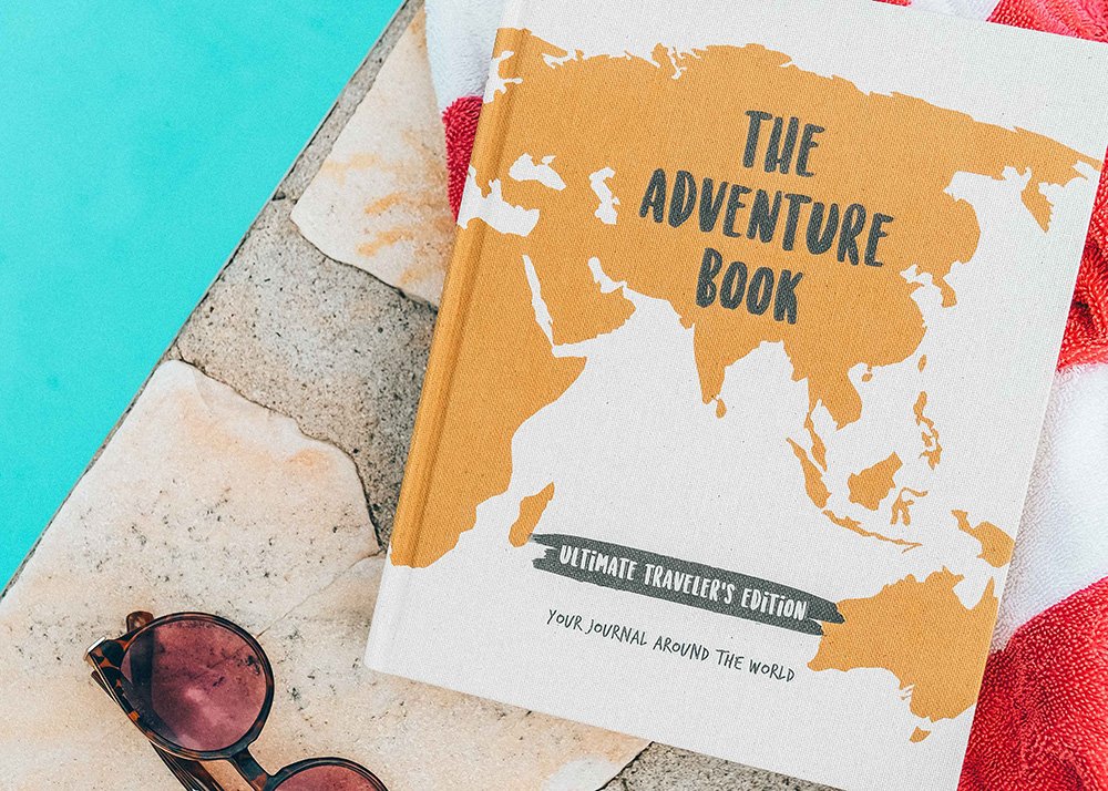 The deluxe white and gold The Adventure Book edition is laying by the side of the pool. A small snippet of the water in the pool is visible as is a pair of sunglasses and a little piece of a red and white towel.