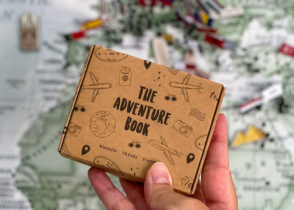A hand holding a small cardboard box labeled "The Adventure Book" with illustrations of travel-related items such as airplanes, cameras, and passports, set against a blurred map background. Inside the box are travel related stamps to scrapbook with.