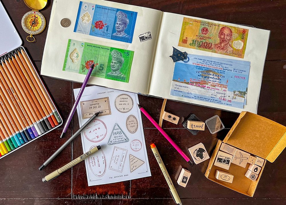 A travel scrapbook lays open on a dark wooden table and is filled with ticket stubs and drawings. Travel related stamps, stickers and brushes and pencils are surrounding the scrapbook
