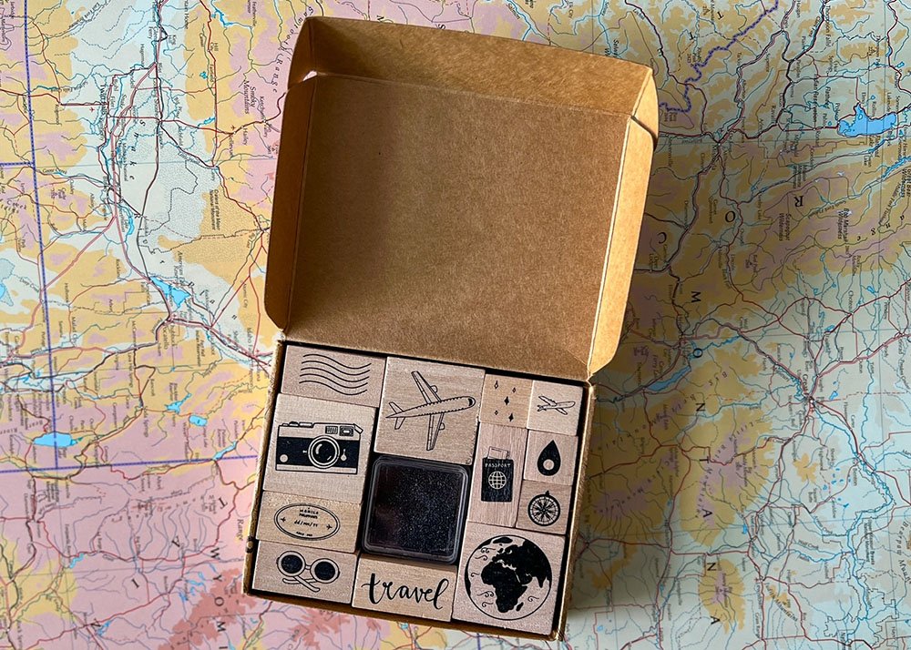 A small carboard box is opened and a little ink box surrounded by 8 travel related stamps are inside. The box is placed on top of a world map