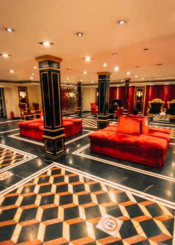 Luxurious cruise ship lobby featuring ornate decor with red couches, black and gold columns, and a geometric-patterned floor.
