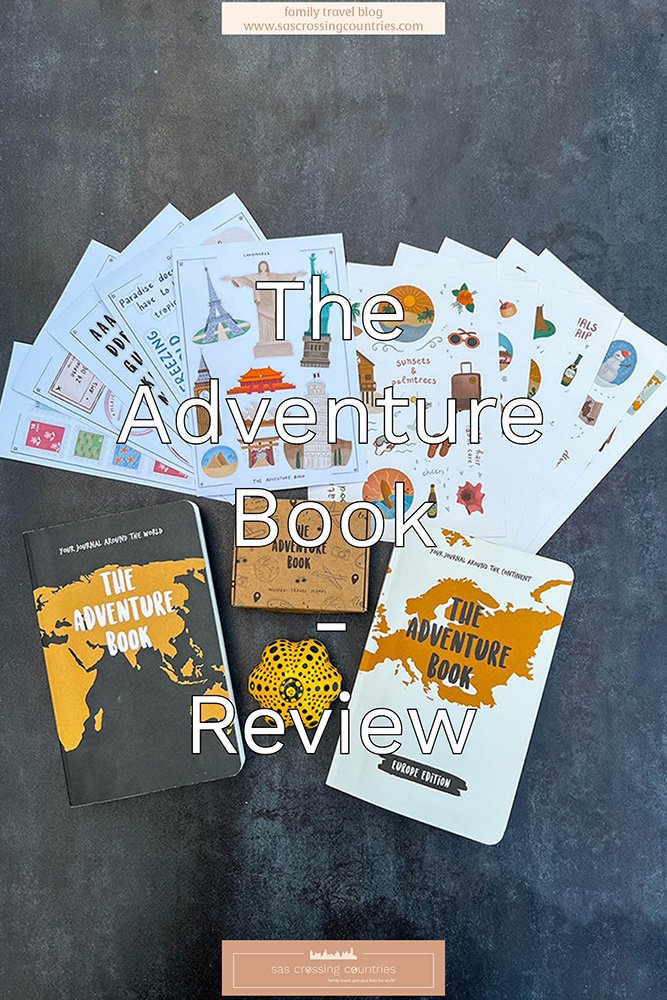 Cover image for a family travel blog post titled "The Adventure Book Review" showcasing a collection of travel-themed stickers and two versions of "The Adventure Book" journals, a black world and a gold and white Europe edition, and a compact cardboard box with travel related stamps inside. The items are arranged on a dark surface with a decorative yellow dice featuring travel icons. The blog URL "www.sascrossingcountries.com" is visible at the top.