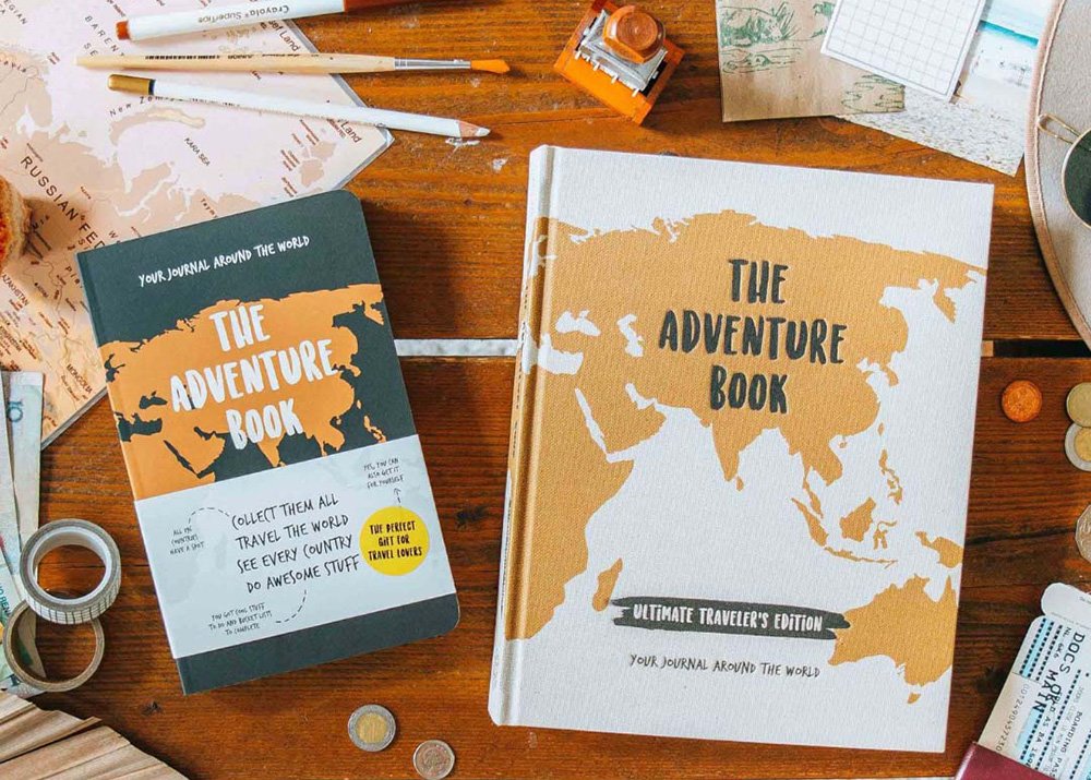 A flat lay image of The Adventure Book - original edition and the new deluxe edition. Both are lying on a brown surface with travel related scrapbook items all around it