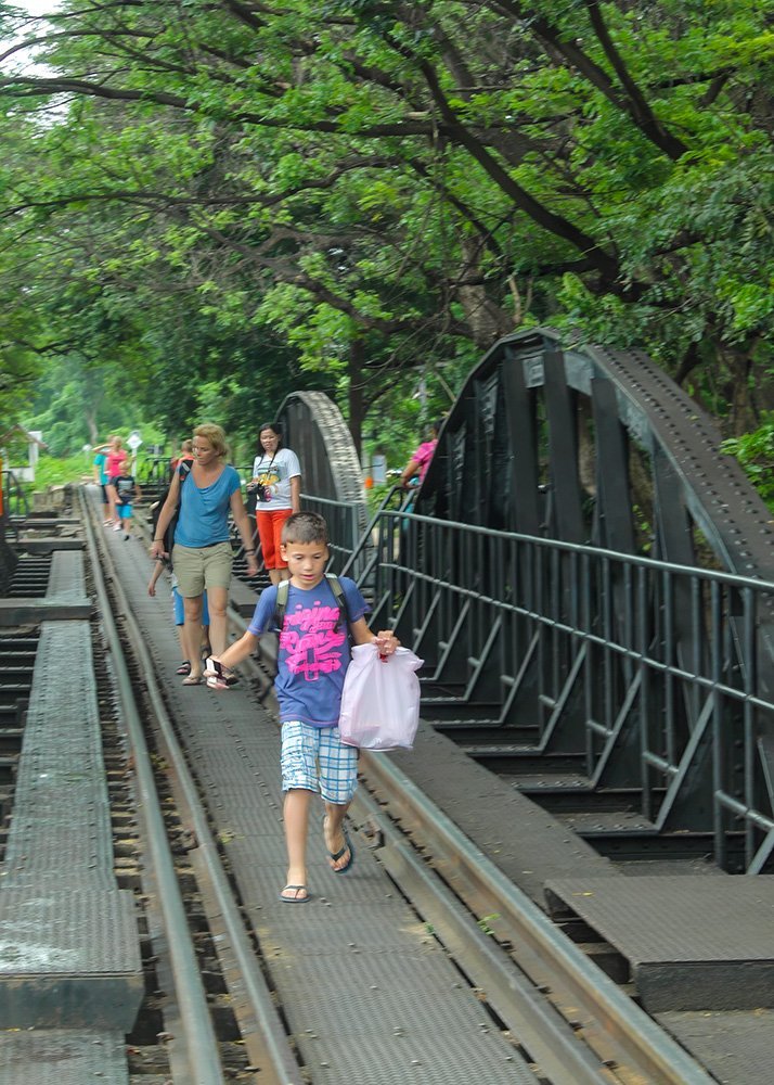 Image of people walking on a bridge: A boy with a plastic bag walks on a metal bridge with other people in the background. The bridge is surrounded by lush green trees.