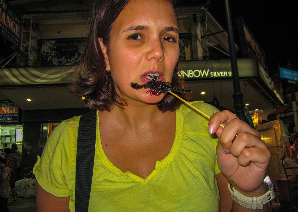Image of a woman eating a scorpion: A woman in a yellow shirt bites into a scorpion on a stick, likely at a night market on Khao San Road, with shops and signs visible in the background.