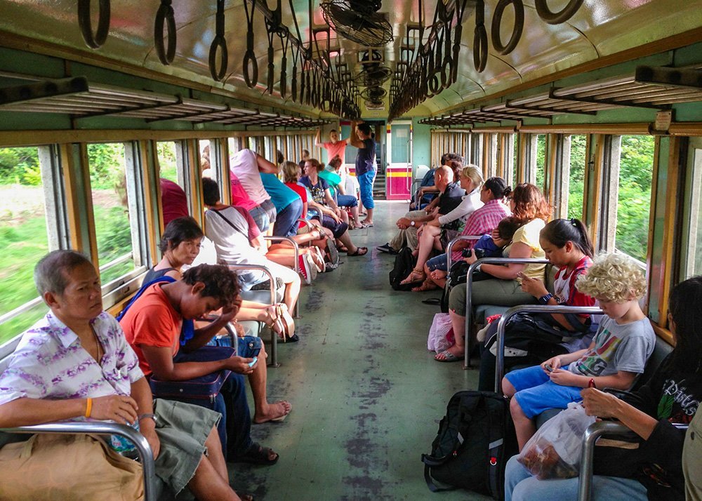 Image of people sitting inside a train: Passengers sit inside a train car in Thailand, some looking out the window and others engaged in various activities. The train interior has metal bars and hanging straps.