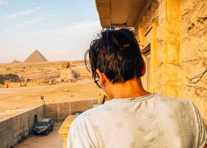 A teenage boy on a balcony overlooking the Sphinx and Pyramids of Giza in the distance.