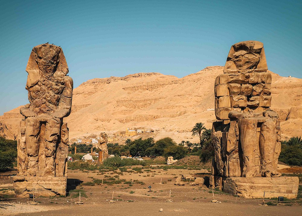 The Colossi of Memnon, two massive stone statues of Pharaoh Amenhotep III, standing in a barren field with desert mountains in the background.