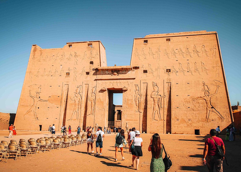 Tourists walking towards the entrance of the Temple of Edfu, with tall walls adorned with carvings.