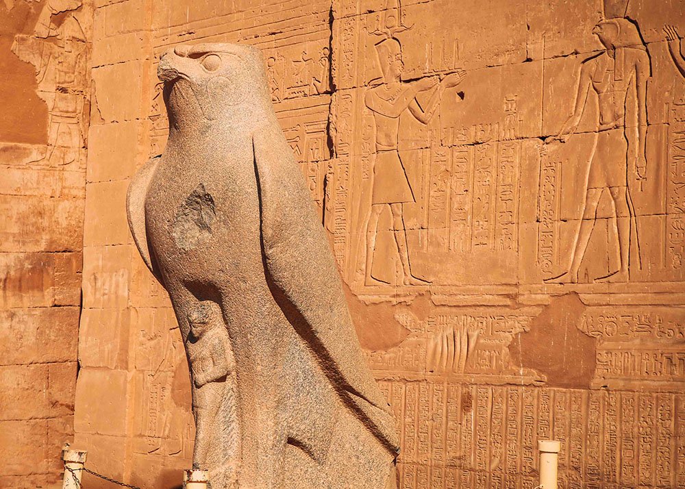 A stone statue of a falcon with hieroglyphics on the wall behind it in the Egyptian temple complex of Edfu