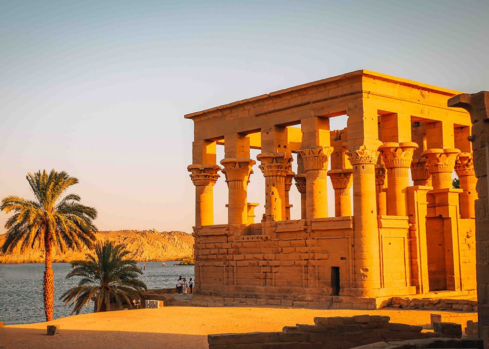 An angle of the Philae Temple complex, featuring a smaller temple with columns and nearby palm trees, overlooking the water.
