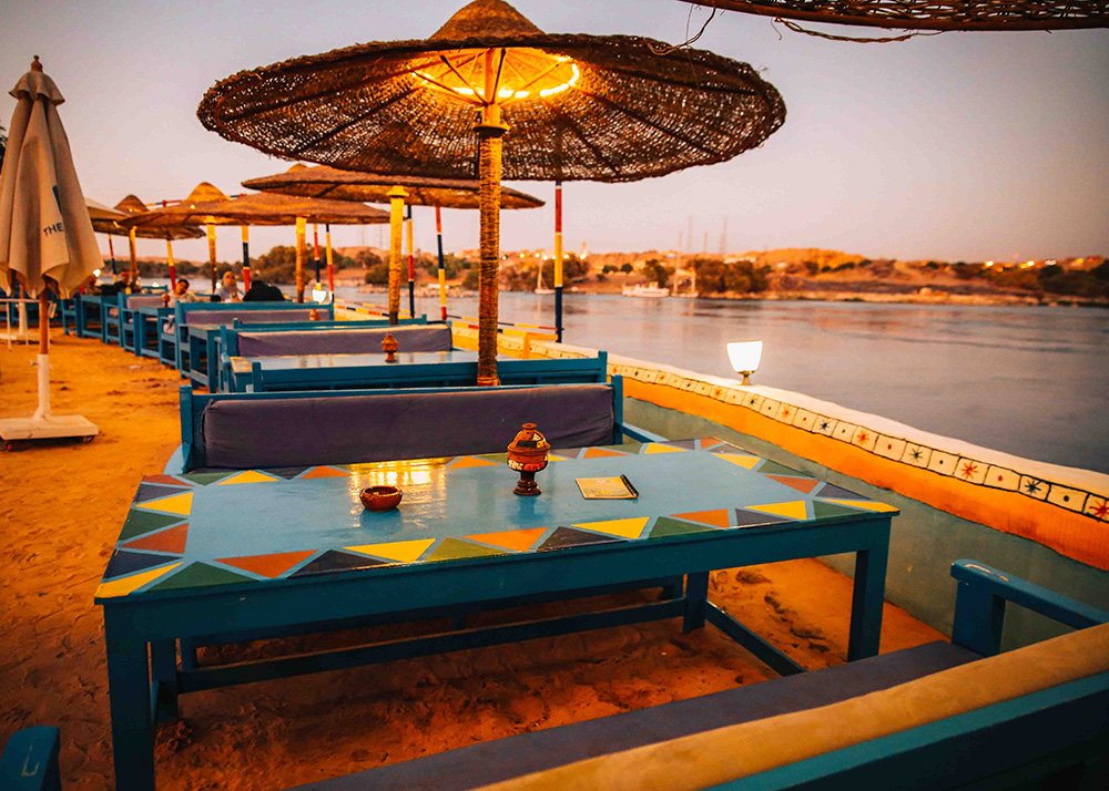 Outdoor dining area with blue tables and benches under large straw umbrellas, by the Nile riverside at dusk.