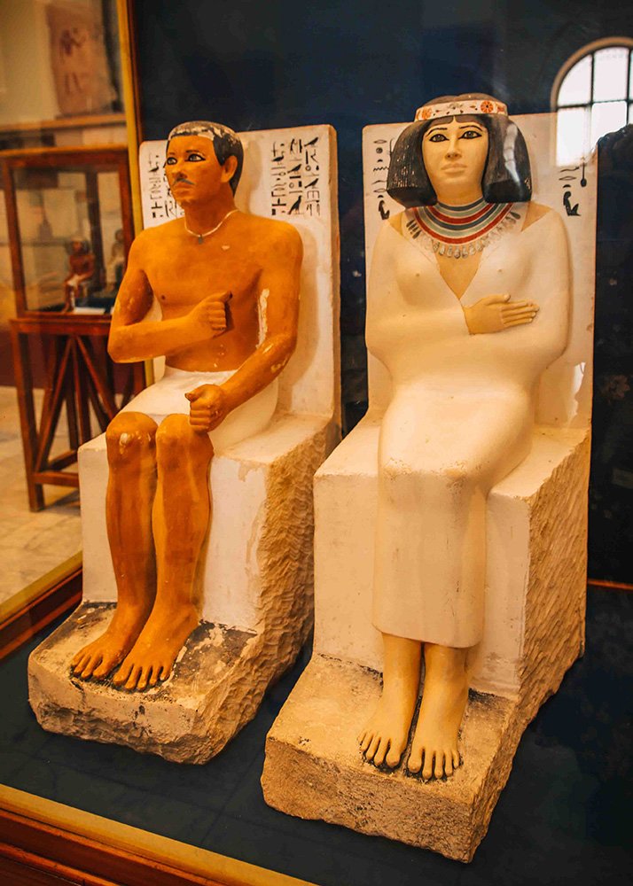 Colorful statues of an ancient Egyptian man and woman seated on stone chairs, with hieroglyphics inscribed behind them.