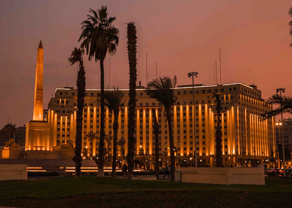 A prominent building with an obelisk and palm trees at Tahrir Square, illuminated by warm lights during twilight.