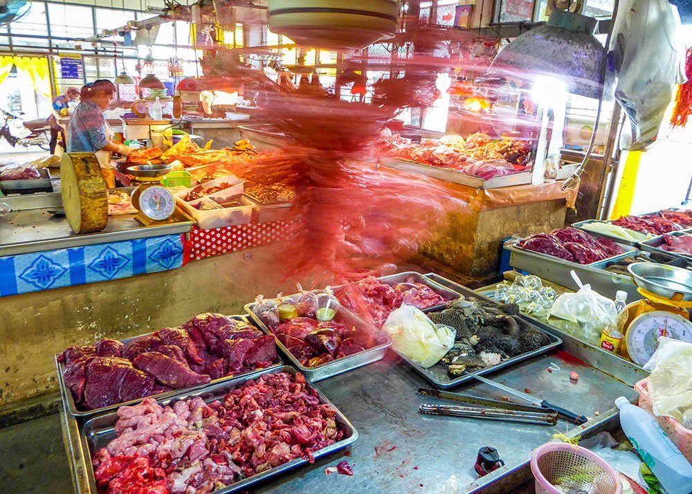 A bustling market stall with various meats on display, with a blur of movement from a vendor slicing meat behind the counter.